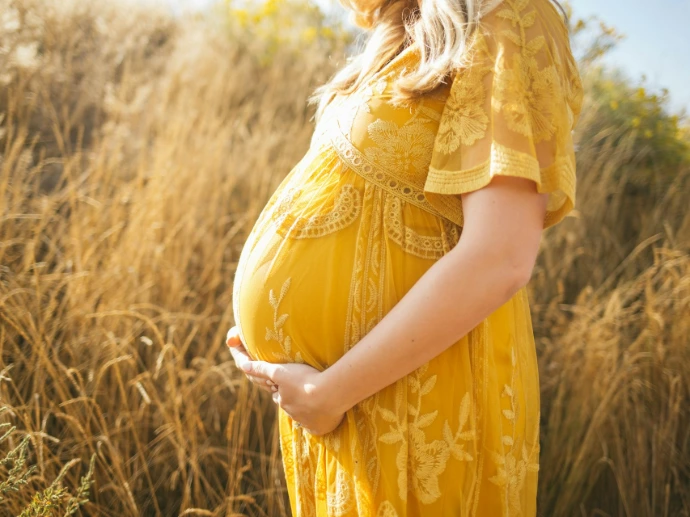 pregnant woman wearing yellow floral dress standing while touching her tummy and facing her right side near brown field during daytime